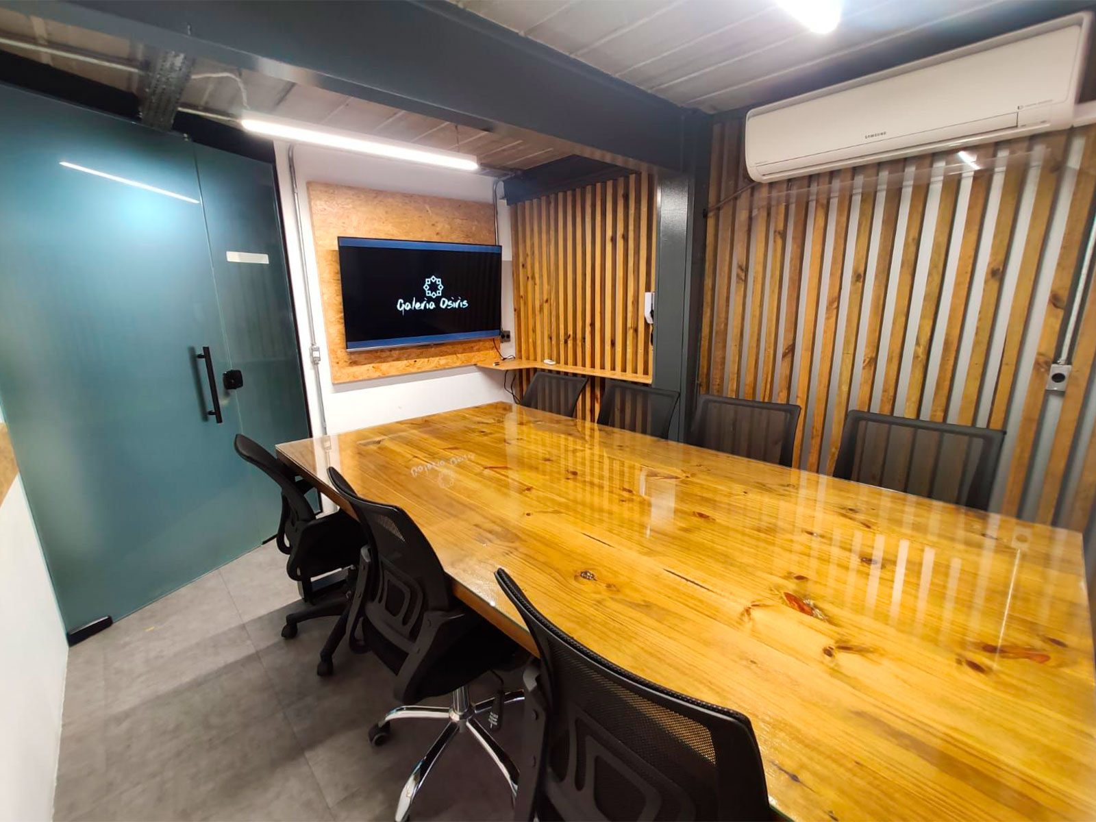 Sala de reunião com mesa de madeira para oito pessoas, cadeiras ergonômicas, TV na parede e ripado de madeira